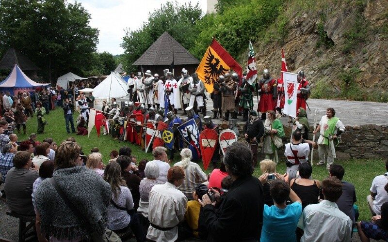 Eine Gruppe von Menschen in mittelalterlichen Kostümen mit Schilden und Fahnen steht im Freien, während eine Menschenmenge vor ihnen sitzt und zusieht.