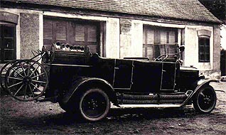 Schwarz-Weiß-Foto eines Autos aus dem frühen 20. Jahrhundert, das vor einem Gebäude mit verschlossenen Fenstern geparkt ist. Auf der Rückseite ist ein Ersatzreifen montiert.