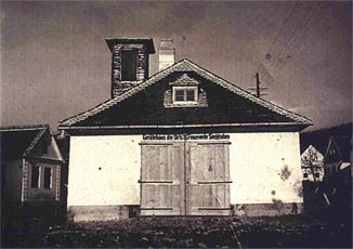 Ein Schwarz-Weiß-Foto eines kleinen, alten Feuerwehrgebäudes mit doppelten Holztüren und einem Glockenturm über dem Eingang.