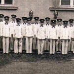 Eine Gruppe von Jungen und zwei erwachsene Männer in Uniform und mit Mützen stehen in einer Reihe vor einem Gebäude mit Fenstern.