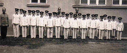 Eine Gruppe von Jungen und zwei erwachsene Männer in Uniform und mit Mützen stehen in einer Reihe vor einem Gebäude mit Fenstern.
