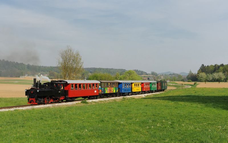 Ein kleiner Dampfzug mit bunten Waggons fährt an einem klaren Tag durch eine grüne, ländliche Landschaft.