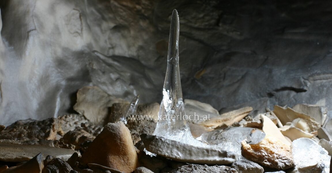 Ein klarer Eisstalagmit erhebt sich aus dem felsigen Höhlenboden, umgeben von verstreuten Steinen und schwach beleuchteten Höhlenwänden.