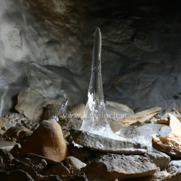 Ein klarer Eisstalagmit erhebt sich aus dem felsigen Höhlenboden, umgeben von verstreuten Steinen und schwach beleuchteten Höhlenwänden.