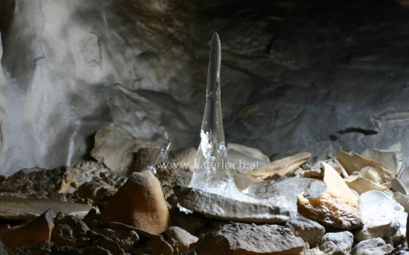 Ein klarer Eisstalagmit erhebt sich aus dem felsigen Höhlenboden, umgeben von verstreuten Steinen und schwach beleuchteten Höhlenwänden.
