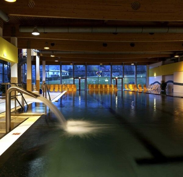 Hallenbad mit Wasserspeier, Metallgeländer und Reihen von gelben Liegestühlen entlang großer Fenster, die Tageslicht hereinlassen.