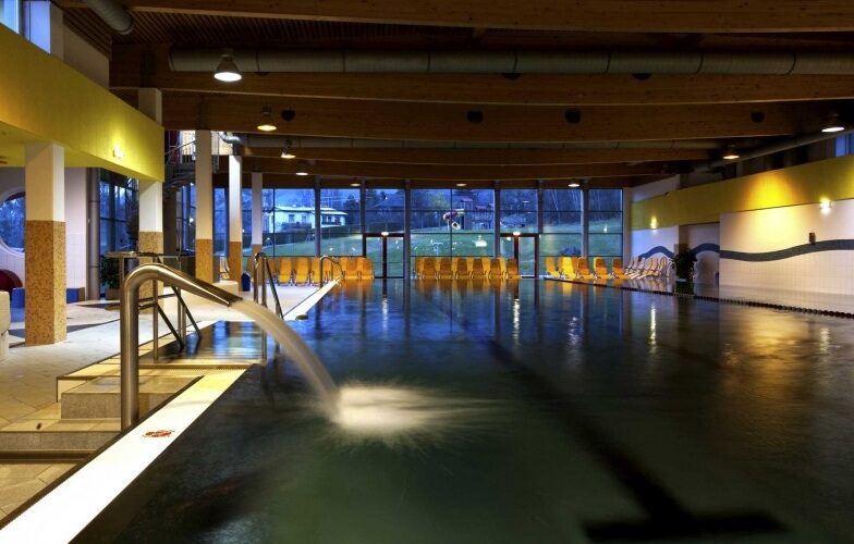 Hallenbad mit Wasserspeier, Metallgeländer und Reihen von gelben Liegestühlen entlang großer Fenster, die Tageslicht hereinlassen.