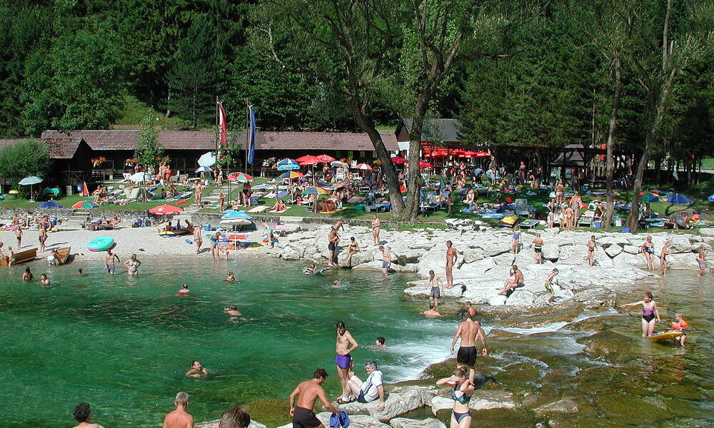 Die Menschen schwimmen, sonnen sich und entspannen sich in der Nähe eines felsigen Ufers und klaren grünen Wassers, mit Bäumen und einer Grasfläche mit Sonnenschirmen und Gebäuden im Hintergrund.