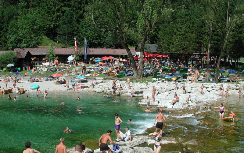 Die Menschen schwimmen, sonnen sich und entspannen sich in der Nähe eines felsigen Ufers und klaren grünen Wassers, mit Bäumen und einer Grasfläche mit Sonnenschirmen und Gebäuden im Hintergrund.