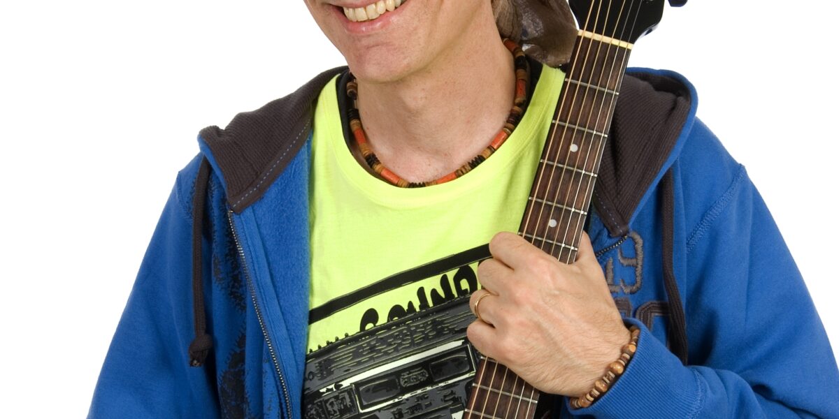 Eine Person, die ein Kopftuch, einen blauen Kapuzenpulli und ein gelbes Hemd trägt, lächelt, während sie eine schwarze Akustikgitarre vor einem weißen Hintergrund hält.