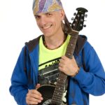 Eine Person, die ein Kopftuch, einen blauen Kapuzenpulli und ein gelbes Hemd trägt, lächelt, während sie eine schwarze Akustikgitarre vor einem weißen Hintergrund hält.