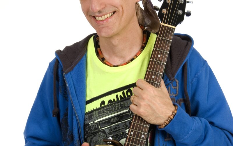 Eine Person, die ein Kopftuch, einen blauen Kapuzenpulli und ein gelbes Hemd trägt, lächelt, während sie eine schwarze Akustikgitarre vor einem weißen Hintergrund hält.