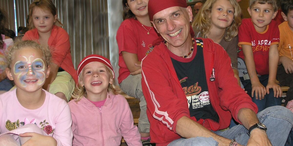 Eine Gruppe von Kindern und ein Erwachsener, einige lächelnd, sitzen in einem Haus zusammen; ein Kind ist geschminkt, ein anderes trägt einen roten Hut.
