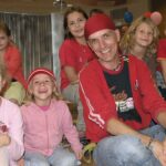 Eine Gruppe von Kindern und ein Erwachsener, einige lächelnd, sitzen in einem Haus zusammen; ein Kind ist geschminkt, ein anderes trägt einen roten Hut.