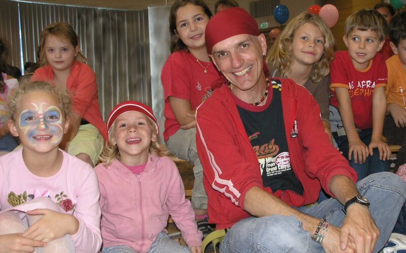 Eine Gruppe von Kindern und ein Erwachsener, einige lächelnd, sitzen in einem Haus zusammen; ein Kind ist geschminkt, ein anderes trägt einen roten Hut.