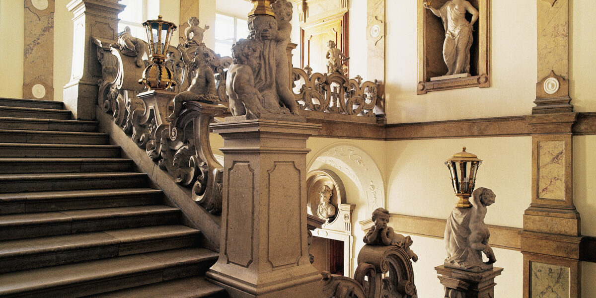 Große Steintreppe mit kunstvollem Geländer, Statuen und Laternen in einem eleganten, historischen Interieur mit Marmorböden und klassischen Wandskulpturen.