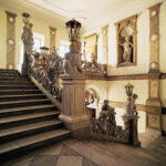 Große Steintreppe mit kunstvollem Geländer, Statuen und Laternen in einem eleganten, historischen Interieur mit Marmorböden und klassischen Wandskulpturen.