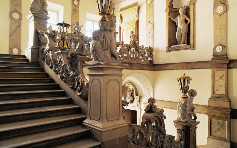 Große Steintreppe mit kunstvollem Geländer, Statuen und Laternen in einem eleganten, historischen Interieur mit Marmorböden und klassischen Wandskulpturen.