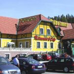 Ein gelb-grünes Restaurant mit roten Dachziegeln, Außentreppen und parkenden Autos auf dem Parkplatz an einem klaren Tag, umgeben von Grün und Bäumen.