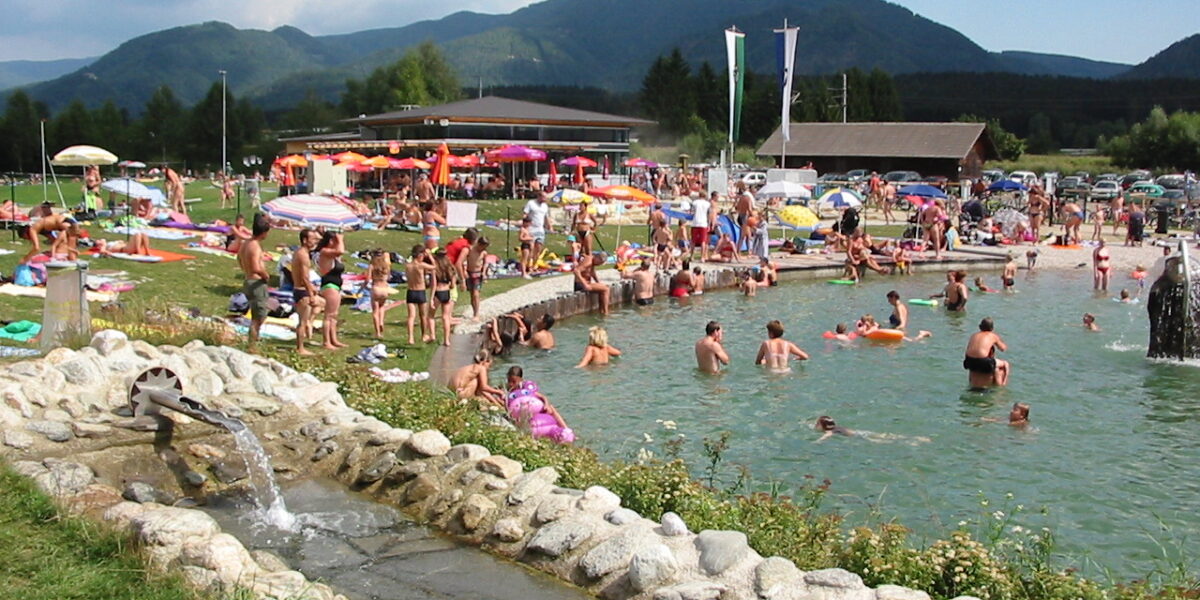 Ein überfüllter Freibadbereich mit Menschen, die sich sonnen, sitzen und schwimmen, umgeben von Gras, Sonnenschirmen und Bergen im Hintergrund.