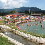 Ein überfüllter Freibadbereich mit Menschen, die sich sonnen, sitzen und schwimmen, umgeben von Gras, Sonnenschirmen und Bergen im Hintergrund.