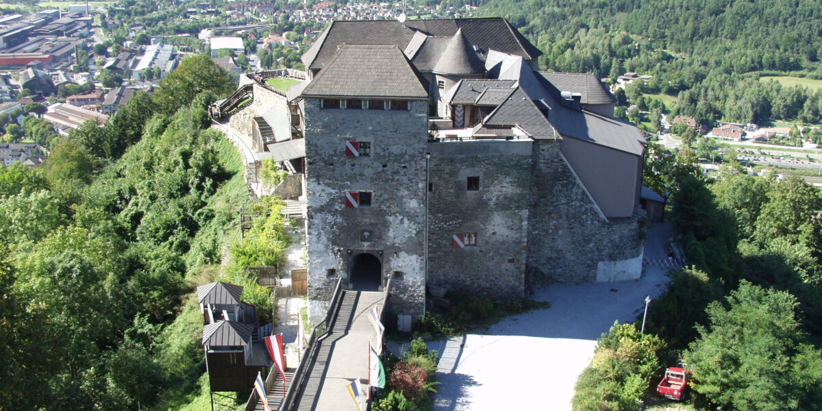 Luftaufnahme einer Steinburg mit mehreren Dächern auf einem Hügel, umgeben von Bäumen und mit Blick auf ein Tal mit Gebäuden und grünen Bergen im Hintergrund.