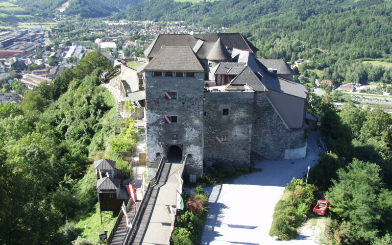 Luftaufnahme einer Steinburg mit mehreren Dächern auf einem Hügel, umgeben von Bäumen und mit Blick auf ein Tal mit Gebäuden und grünen Bergen im Hintergrund.