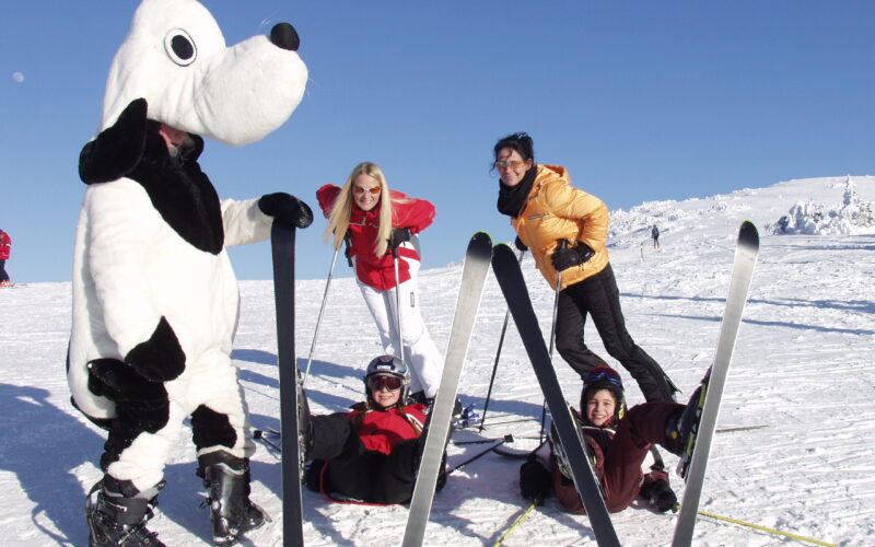 Vier Personen posieren auf einer verschneiten Piste mit einer Person in einem Hundemaskottchen-Kostüm; zwei stehen, während zwei mit V-förmig angeordneten Skiern auf dem Schnee sitzen.