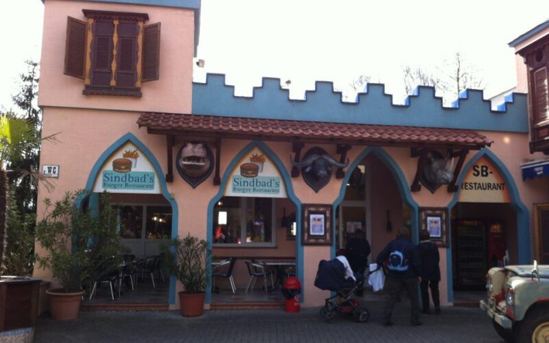 Ein Restaurant mit einer schlossähnlichen Fassade, gewölbten Eingängen und Schildern mit der Aufschrift "Sindbad's Burger Restaurant". In der Nähe des Eingangs stehen Menschen mit einem Kinderwagen.