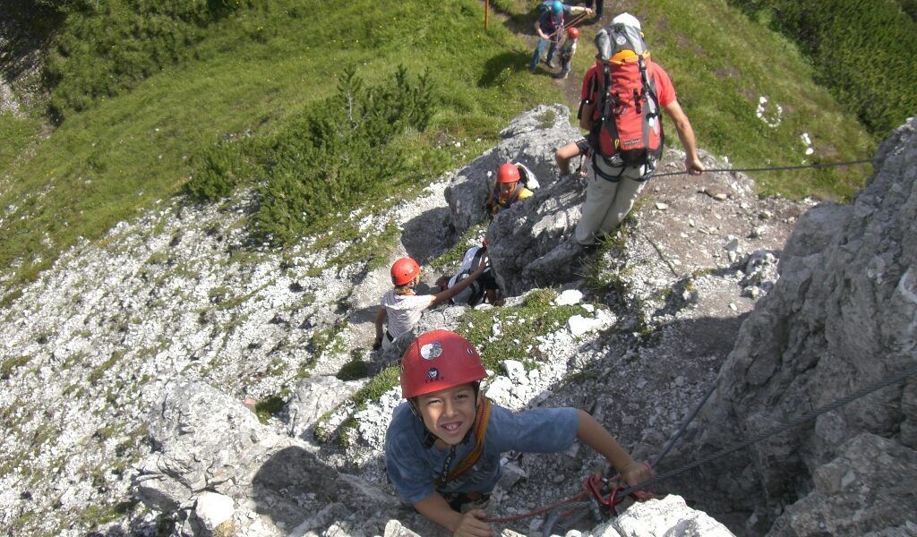 Eine Gruppe von Kletterern mit Helmen und Klettergurten steigt an einem sonnigen Tag mit Hilfe von Sicherungsseilen einen felsigen, steilen Hang hinauf.