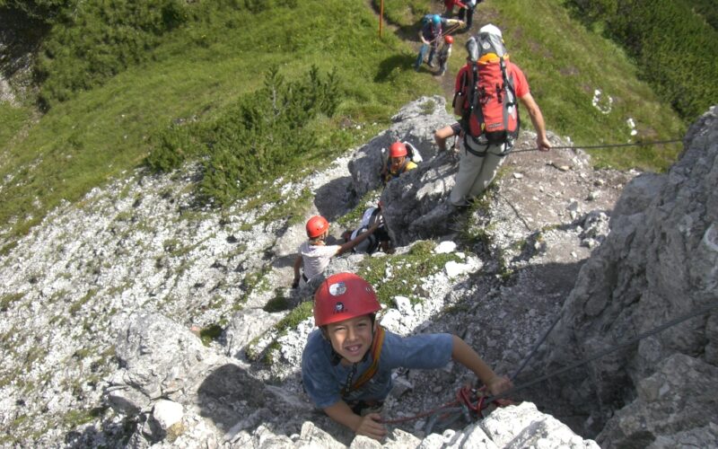 Eine Gruppe von Kletterern mit Helmen und Klettergurten steigt an einem sonnigen Tag mit Hilfe von Sicherungsseilen einen felsigen, steilen Hang hinauf.