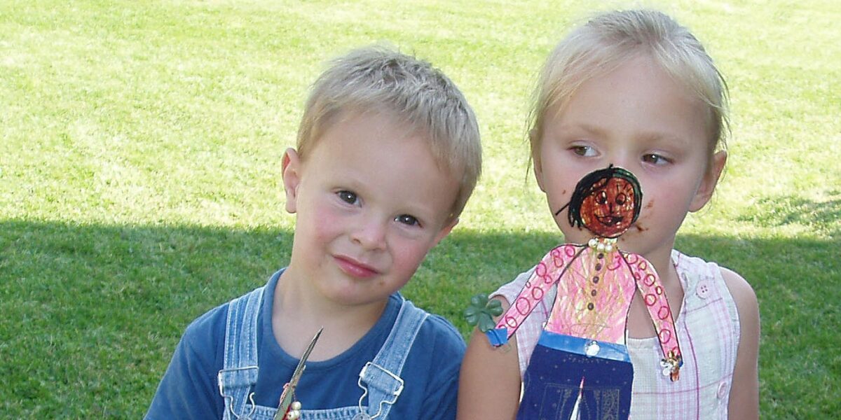 Ein kleiner Junge in einem Overall und ein kleines Mädchen mit einer handgefertigten Papierpuppe stehen zusammen auf einer Wiese.