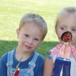 Ein kleiner Junge in einem Overall und ein kleines Mädchen mit einer handgefertigten Papierpuppe stehen zusammen auf einer Wiese.