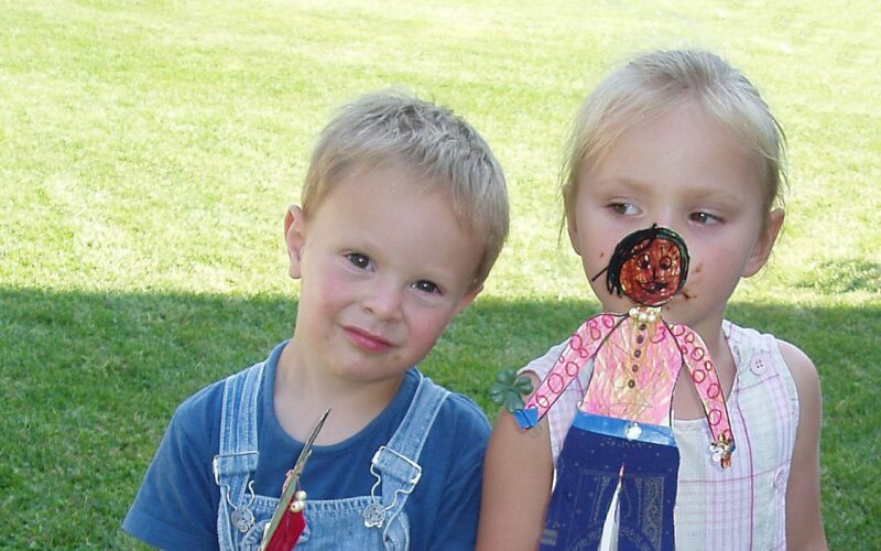 Ein kleiner Junge in einem Overall und ein kleines Mädchen mit einer handgefertigten Papierpuppe stehen zusammen auf einer Wiese.