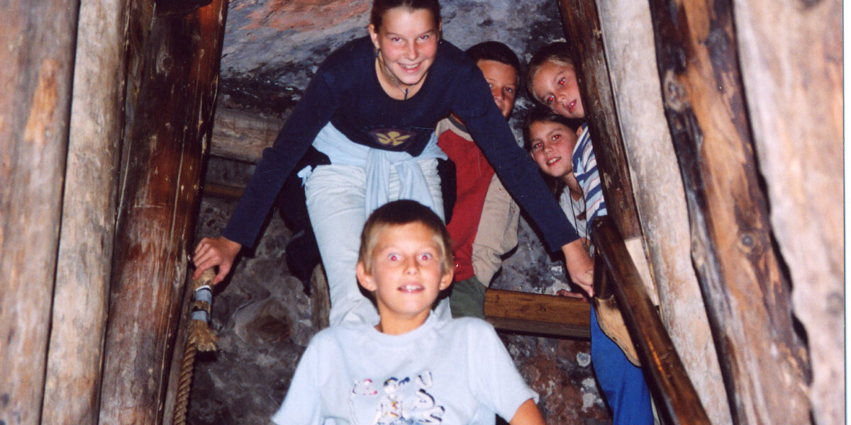 Fünf Kinder posieren gemeinsam auf einer Holztreppe in einem Tunnel oder einer Höhle, die von Holzbalken umgeben zu sein scheint.