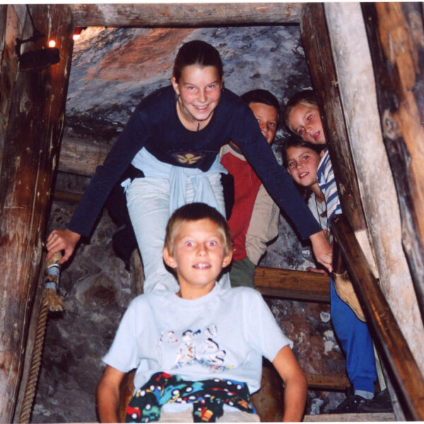 Fünf Kinder posieren gemeinsam auf einer Holztreppe in einem Tunnel oder einer Höhle, die von Holzbalken umgeben zu sein scheint.