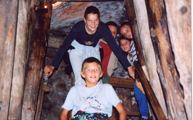 Fünf Kinder posieren gemeinsam auf einer Holztreppe in einem Tunnel oder einer Höhle, die von Holzbalken umgeben zu sein scheint.