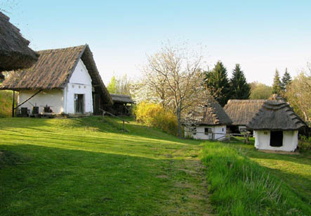 Mehrere weiße, strohgedeckte Häuser stehen in einer grasbewachsenen, hügeligen Landschaft mit Bäumen und klarem Himmel im Hintergrund.