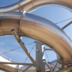 Gebogene, glänzende Metallrohre sind miteinander verbunden und werden von Streben vor einem klaren blauen Himmel gestützt.