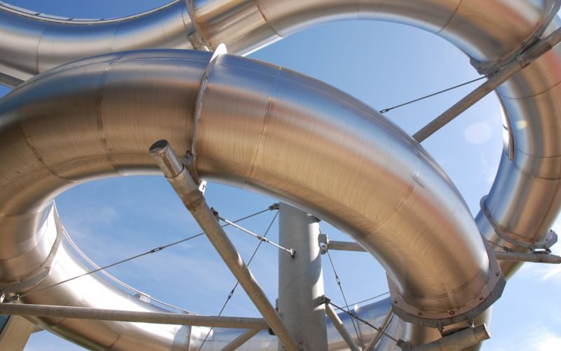 Gebogene, glänzende Metallrohre sind miteinander verbunden und werden von Streben vor einem klaren blauen Himmel gestützt.