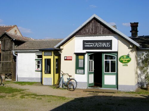 Kleines ländliches Gebäude mit einem „Gasthaus“-Schild, einer gelben Telefonzelle, einem davor geparkten Fahrrad und einem Bitburger-Bier-Schild an der Wand.