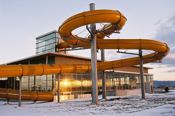 Eine große gelbe Wasserrutsche im Freien windet sich um Metallstützen neben einem modernen Glasgebäude, während der Boden mit Schnee bedeckt ist und die Sonne sich in den Fenstern spiegelt.