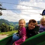 Drei kleine Kinder rutschen auf einer grünen Rutsche im Freien mit Bergen, Bäumen und einer Seilbahn im Hintergrund unter einem teilweise bewölkten Himmel hinunter.
