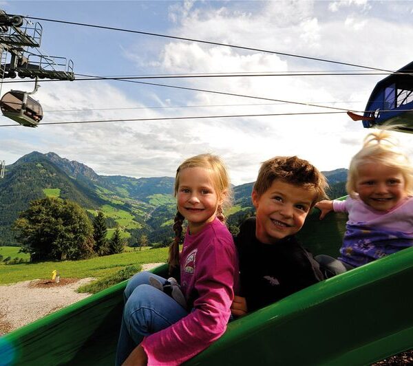 Drei kleine Kinder rutschen auf einer grünen Rutsche im Freien mit Bergen, Bäumen und einer Seilbahn im Hintergrund unter einem teilweise bewölkten Himmel hinunter.