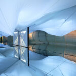 Raum mit verspiegeltem Boden, weißer Stoffdecke, Holzwand mit Tür und einem vertikalen Landschaftsfoto, das sich im Spiegel spiegelt.