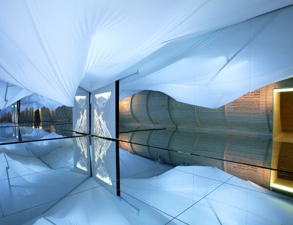 Raum mit verspiegeltem Boden, weißer Stoffdecke, Holzwand mit Tür und einem vertikalen Landschaftsfoto, das sich im Spiegel spiegelt.