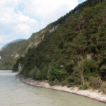 Ein Fluss fließt entlang eines bewaldeten Hügels bei teilweise bewölktem Himmel, im Hintergrund sind Berge zu sehen.