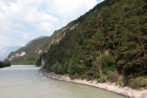 Ein Fluss fließt entlang eines bewaldeten Hügels bei teilweise bewölktem Himmel, im Hintergrund sind Berge zu sehen.