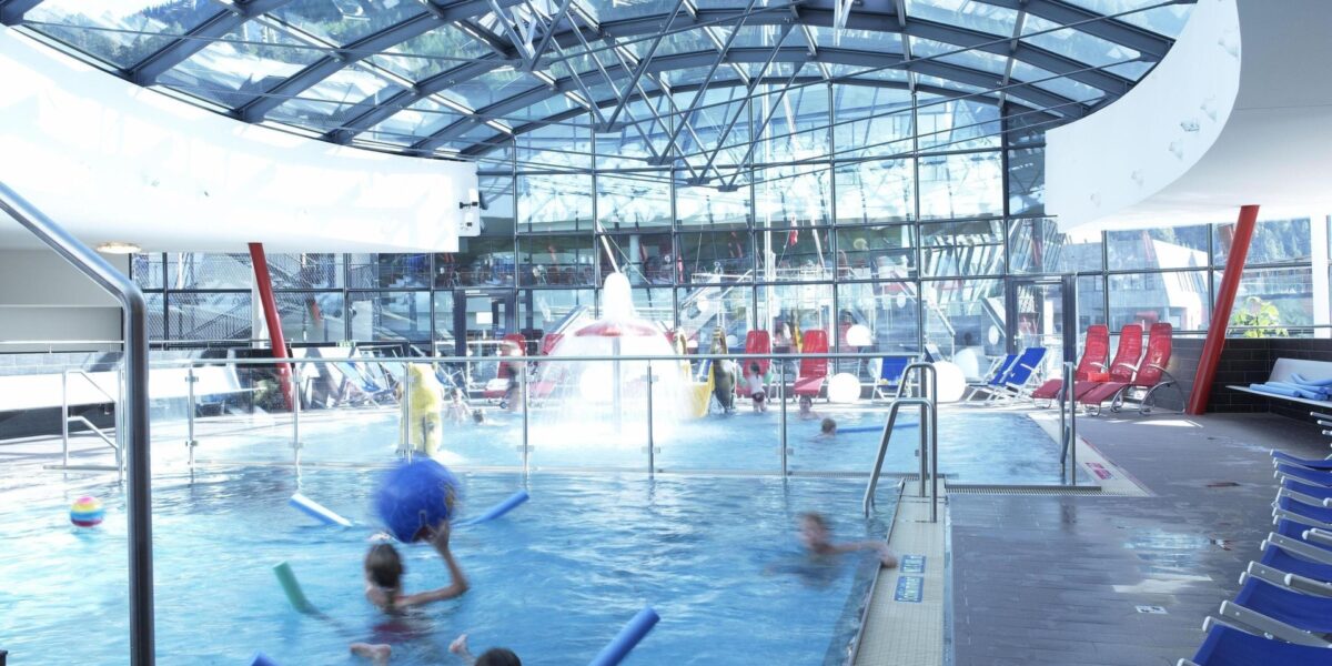 Innenpool mit im Wasser spielenden Kindern, umgeben von Liegestühlen und umschlossen von einem Glasdach und Glaswänden.