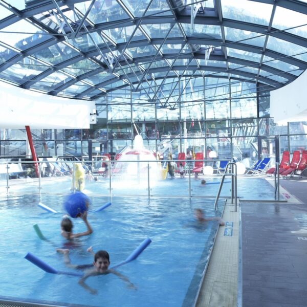 Innenpool mit im Wasser spielenden Kindern, umgeben von Liegestühlen und umschlossen von einem Glasdach und Glaswänden.